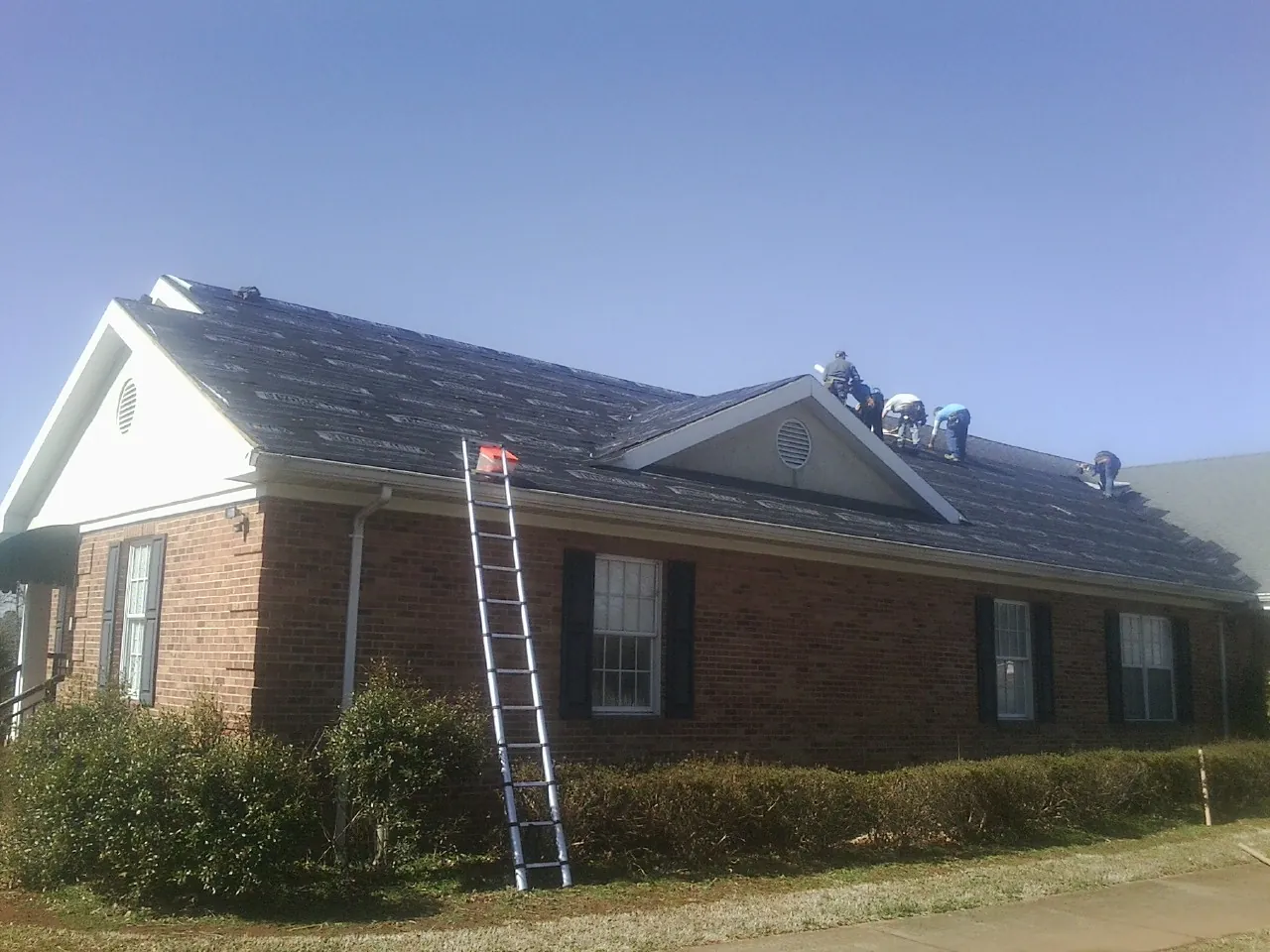 Crew tearing shingles off a brick ranch roof with a ladder set — Metal Roof Repair work by our crew in Lake Orion
