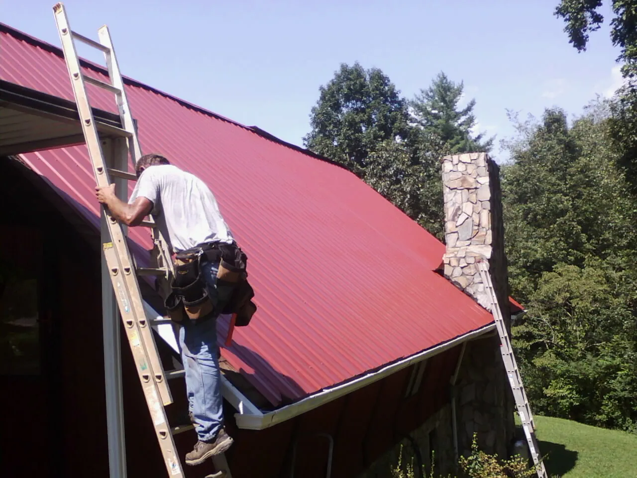 Roofer on a ladder beside a red metal roof and stone chimney — Soffit & Fascia Repair by Roofing Brothers in Lake Orion
