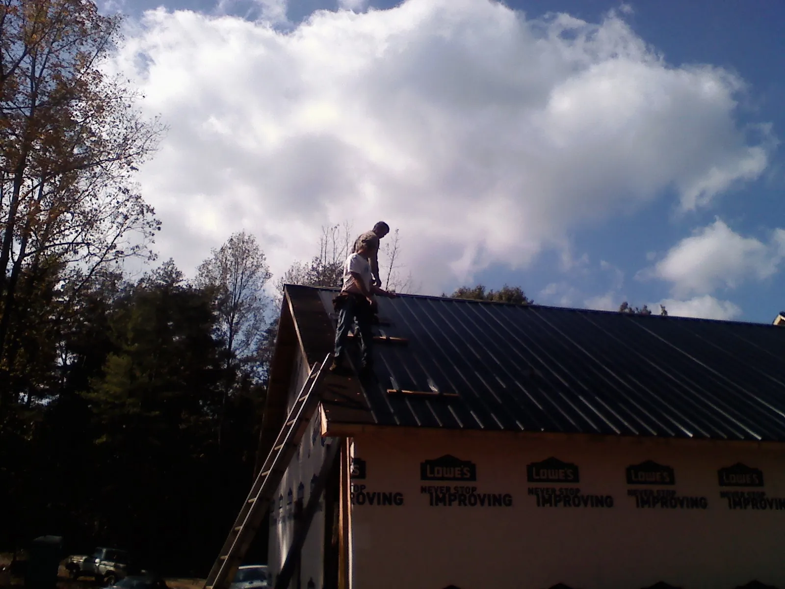 Roofer fastening dark metal panels high against a cloudy sky, from a Roof Inspection job in Lake Orion